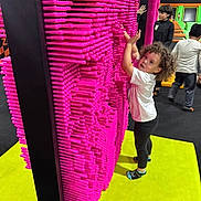 Mariah is registered to the contest to win money with this photo: child, curly_hair, pin_art_board, pink, yellow_floor, play_area, indoor, people, kids, black_pants, white_shirt, socks, curious, interactive_toy, fun, play, colorful, standing, hands_up, portrait