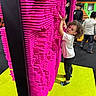 child, curly_hair, pin_art_board, pink, yellow_floor, play_area, indoor, people, kids, black_pants, white_shirt, socks, curious, interactive_toy, fun, play, colorful, standing, hands_up, portrait
