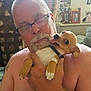 man, dog, glasses, shirtless, indoor, holding, pet, happy, small_dog, beard, tattoo, plant, photo_frames, couch, furniture, close_up, portrait, smiling_dog, home, cozy
