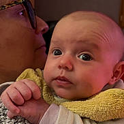 Elena participe au concours pour gagner de l'argent avec cette photo : baby, infant, adult, glasses, yellow_bib, hand, face, indoor, warm_lighting, portrait, closeup, person, holding, curious, skin, clothing, head, eyes, expression, cozy