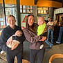 Finlay is registered to the contest to win money with this photo: baby, cafe, casual_clothing, child, group, holding_baby, hoodie, indoor, people, person, portrait, restaurant, sneakers, social_gathering, standing, stone_wall, two_women, wood_floor, yellow_wall, young_adult