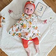 Louise a rejoint le concours — aidez-le/la à gagner de superbes lots ! baby, child, clothing, cute, face, floral_dress, hand, head, headband, indoors, infant, legs, lying_down, milestone_card, orange_leggings, person, smiling, soft_texture, white_blanket, wooden_floor