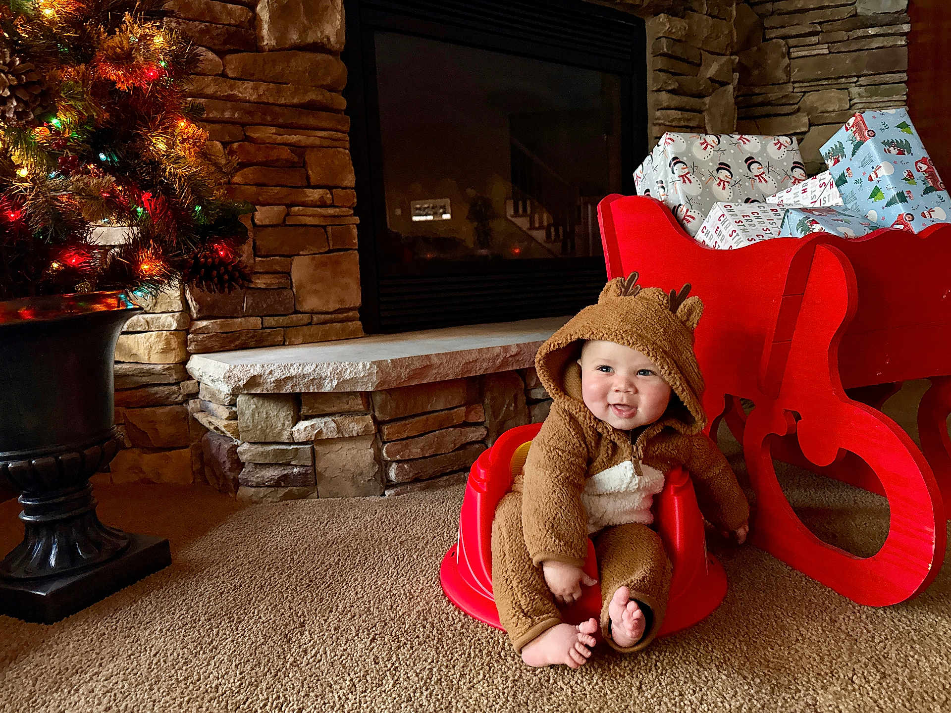 Nickeil joined the competition — help win amazing prizes! baby, infant, reindeer_onesie, red_baby_seat, christmas_sleigh, wrapped_gifts, fireplace, stone_wall, carpet, christmas_tree, holiday_lights, festive, smiling, cute, indoors, cozy, child, holiday_decor, seasonal, happy