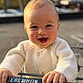 baby, child, smile, happy, shopping_cart, knitted_sweater, hand, face, outdoor, parking_lot, daylight, person, clothing, blond_hair, cute, closeup, portrait, retail, infant, holding
