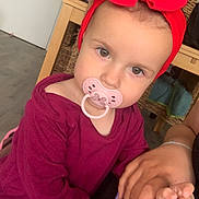 Ruby a rejoint le concours — aidez-le/la à gagner de superbes lots ! toddler, child, pacifier, headband, red_bow, holding_hands, indoor, furniture, wicker_chair, wooden_table, purple_clothing, person, skin, face, eyes, hair, hand, nail_polish, floor, curious_expression