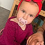 toddler, child, pacifier, headband, red_bow, holding_hands, indoor, furniture, wicker_chair, wooden_table, purple_clothing, person, skin, face, eyes, hair, hand, nail_polish, floor, curious_expression