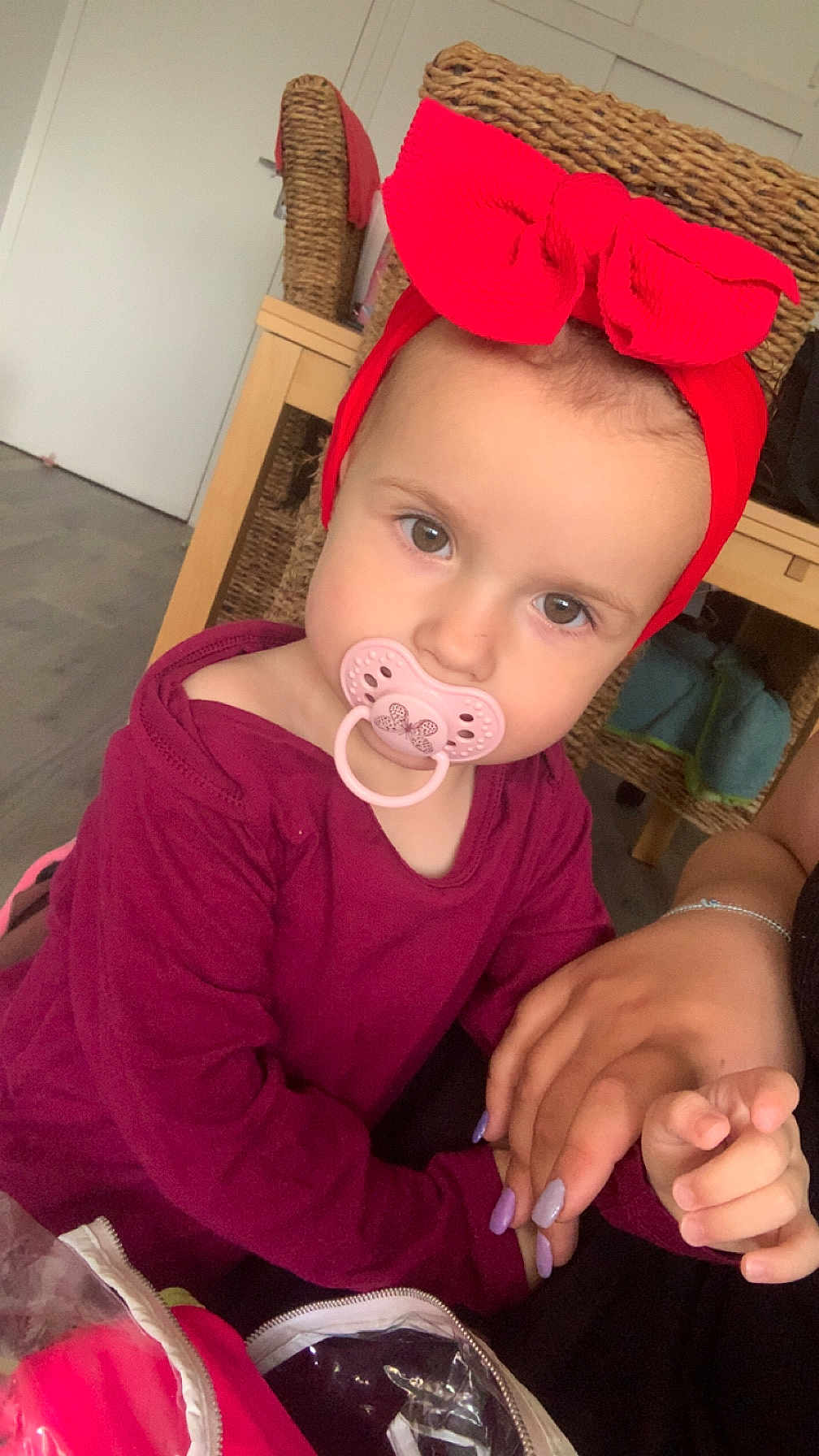 Ruby a rejoint le concours — aidez-le/la à gagner de superbes lots ! toddler, child, pacifier, headband, red_bow, holding_hands, indoor, furniture, wicker_chair, wooden_table, purple_clothing, person, skin, face, eyes, hair, hand, nail_polish, floor, curious_expression