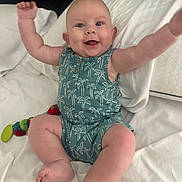 Kai joined the competition — help win amazing prizes! baby, romper, palm_tree_pattern, smiling, arms_up, bed, white_bedding, toy, colorful, happy, infant, cute, chubby, sitting, indoors, child, person, playful, skin, face