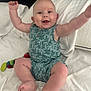 baby, romper, palm_tree_pattern, smiling, arms_up, bed, white_bedding, toy, colorful, happy, infant, cute, chubby, sitting, indoors, child, person, playful, skin, face