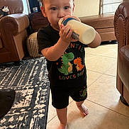 Grayson is registered to the contest to win money with this photo: toddler, child, bottle, drinking, black_clothing, dinosaur_print, barefoot, indoor, living_room, carpet, leather_sofa, tile_floor, cute, person, young_child, hair, furniture, window, natural_light, home