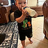 toddler, child, bottle, drinking, black_clothing, dinosaur_print, barefoot, indoor, living_room, carpet, leather_sofa, tile_floor, cute, person, young_child, hair, furniture, window, natural_light, home