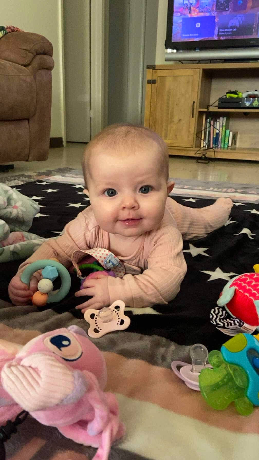 Maisie is registered to the contest to win money with this photo: arm, baby, baby_toddler_clothing, blue, cheek, child, flooring, fun, green, head, iris, mouth, nose, person, room, sitting, skin, stuffed_toy, television, toddler