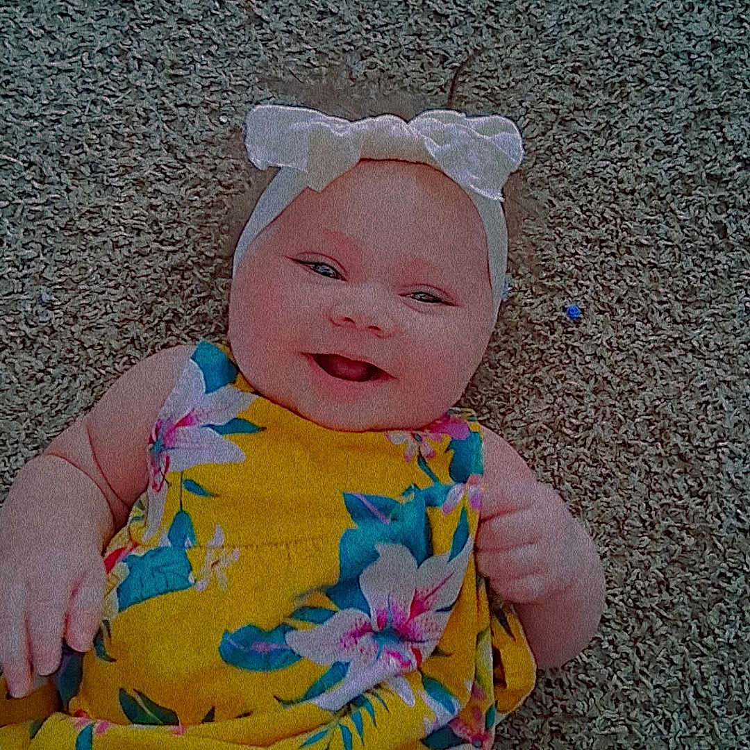 Lilly joined the competition — help win amazing prizes! baby, infant, child, smiling, portrait, indoor, carpet, floral_dress, yellow_clothing, hair_bow, headband, lying_down, happy, face, arms, hands, colorful, text_overlay, floor, photography