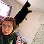 Spooky is registered to the contest to win money with this photo: person, cat, indoor, selfie, glasses, hoodie, jumping_cat, leaping, bed, quilt, door, window, curtain, light, wall, hair, facial_expression, nose_ring, eyewear, feline