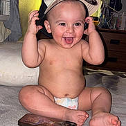 Adam joined the competition — help win amazing prizes! baby, diaper, cap, tongue_out, smiling, indoor, smartphone, television, furniture, couch, blanket, child, cute, happy, playful, person, room, sitting, headwear, infant
