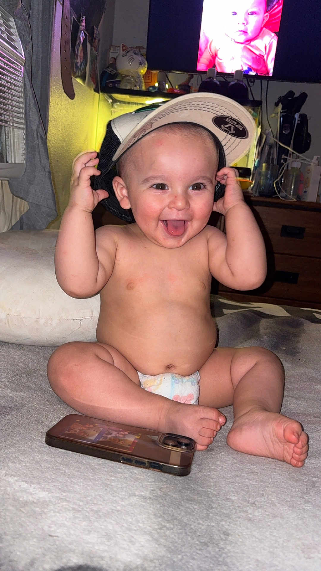 Adam joined the competition — help win amazing prizes! baby, diaper, cap, tongue_out, smiling, indoor, smartphone, television, furniture, couch, blanket, child, cute, happy, playful, person, room, sitting, headwear, infant