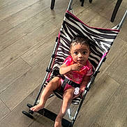 Ariya is registered to the contest to win money with this photo: baby, stroller, zebra_print, pink_clothing, pacifier, wooden_floor, footwear, indoor, child, person, sitting, curious, infant, baby_boy, black_hair, barefoot, seat_belt, toy, flooring, casual