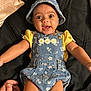 Ariya is registered to the contest to win money with this photo: baby, child, infant, denim, hat, overalls, yellow_shirt, smiling, happy, lying_down, black_background, cute, playful, clothing, person, portrait, indoors, fabric, hand, skin