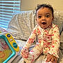 Ariya is registered to the contest to win money with this photo: baby, child, pajamas, floral_pattern, curly_hair, toy, couch, blanket, window_blinds, happy, surprised, indoor, person, infant, sitting, smiling, cozy, home, playtime, cute