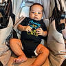 baby, infant, stroller, child, onesie, black_clothing, face, legs, feet, hand, baby_bottle_graphic, text_on_clothing, beige, fabric, seat, comfort, indoor, curious, calm, person