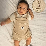 Grabiel Jesus is registered to the contest to win money with this photo: baby, infant, overalls, teddy_bear, blanket, white, lying_down, cute, smiling, celebration, sign, text, socks, child, portrait, indoors, soft_lighting, happy, young, person