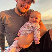 Violet Mae is registered to the contest to win money with this photo: baby, beach, cap, casual_clothing, facial_hair, family, happy, holding, man, nature, ocean, outdoor, pajamas, portrait, sand, sky, smile, sunset, tattoo, warm_light