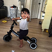 Romina is registered to the contest to win money with this photo: toddler, child, balance_bike, indoor, wooden_floor, laundry_basket, fan, dresser, door, smile, peace_sign, barefoot, white_shirt, black_pants, toy, room, person, happy, floor, home