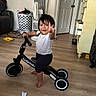 toddler, child, balance_bike, indoor, wooden_floor, laundry_basket, fan, dresser, door, smile, peace_sign, barefoot, white_shirt, black_pants, toy, room, person, happy, floor, home