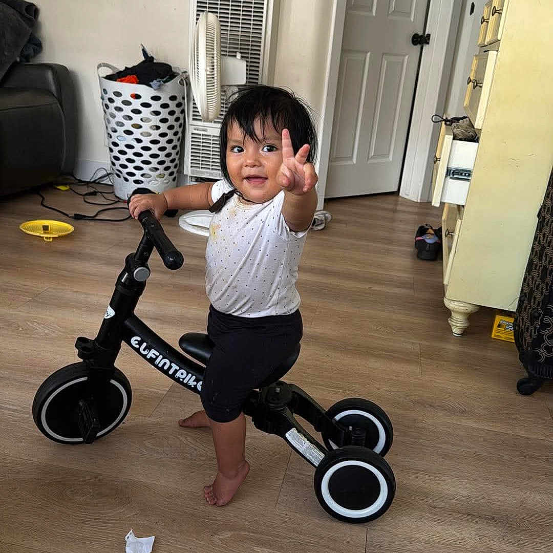 Romina is registered to the contest to win money with this photo: toddler, child, balance_bike, indoor, wooden_floor, laundry_basket, fan, dresser, door, smile, peace_sign, barefoot, white_shirt, black_pants, toy, room, person, happy, floor, home