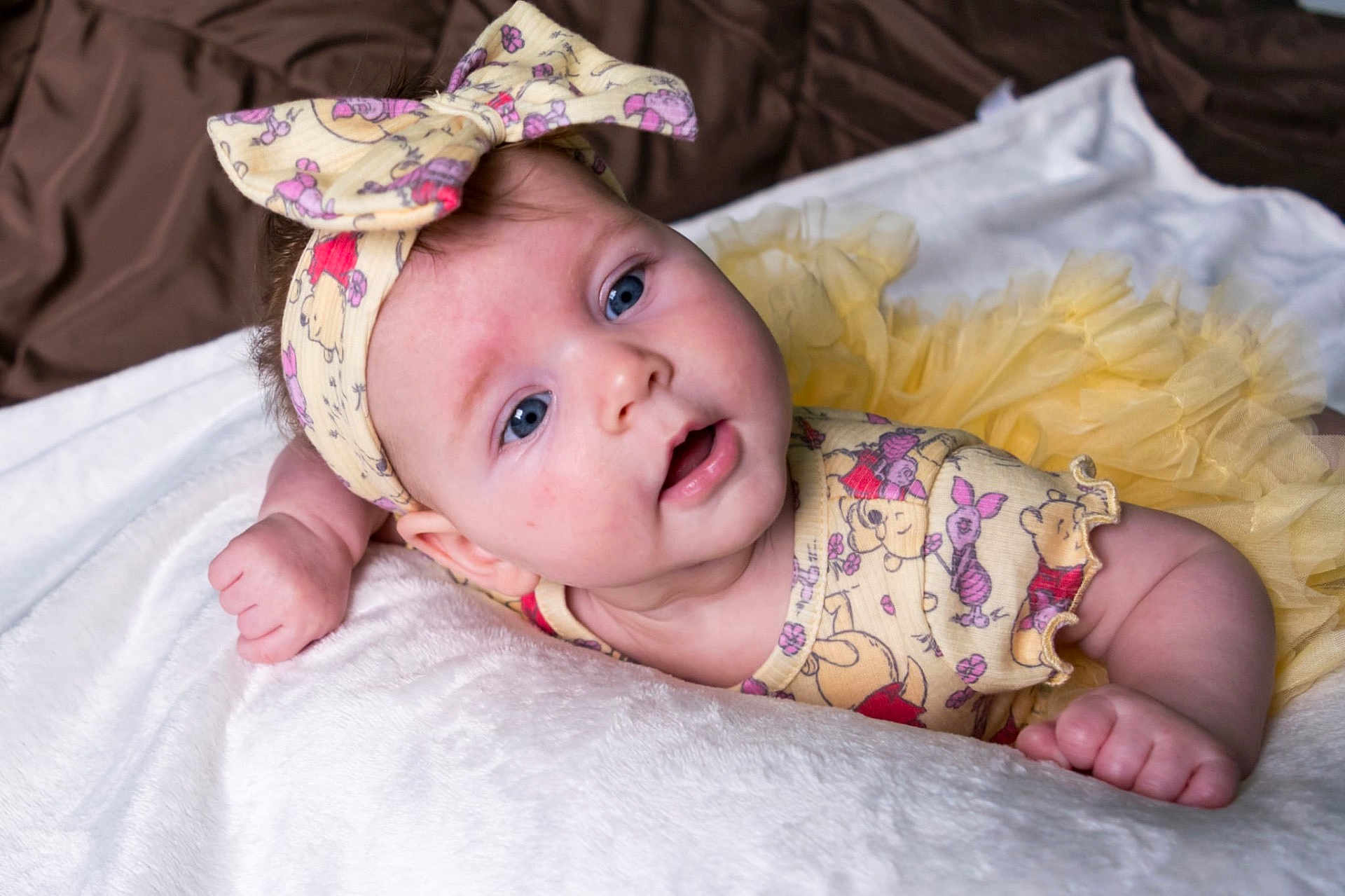 Elaina joined the competition — help win amazing prizes! baby, infant, blue_eyes, headband, bow, yellow_outfit, cartoon_print, tutu, blanket, soft_texture, lying_down, cute, child, portrait, indoors, skin, hand, expression, clothing, person