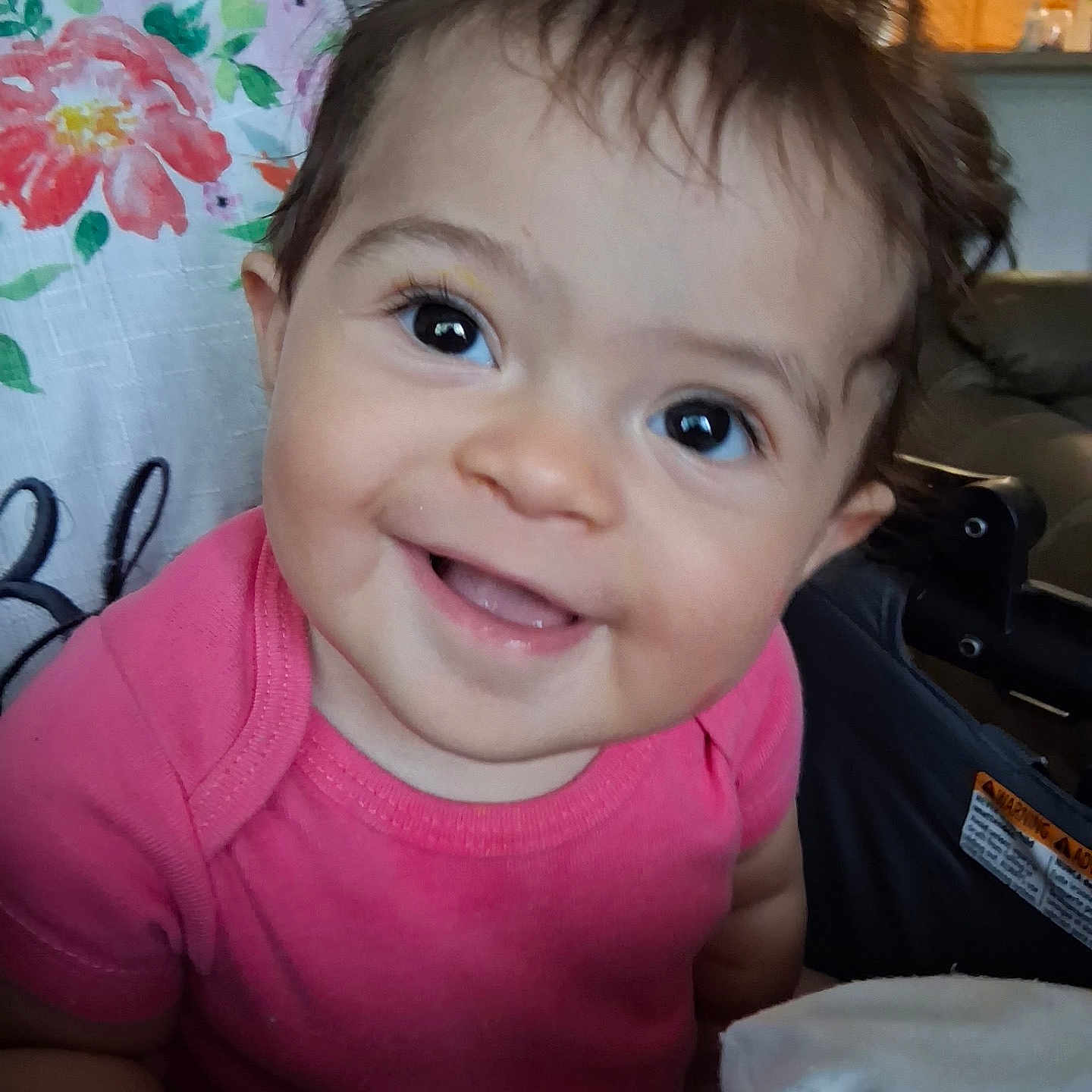 Eliana is registered to the contest to win money with this photo: baby, child, smiling, happy, face, pink_shirt, indoors, portrait, cute, curly_hair, eyes, infant, closeup, expression, person, home, pillow, fabric, seat, cozy
