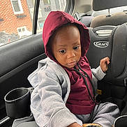 Tymir is registered to the contest to win money with this photo: toddler, child, car_seat, hoodie, gray_sweatpants, shoe, car_interior, window, brick_building, rain, seatbelt, cup_holder, curious, casual_clothing, indoors, portrait, young_child, seat, person, looking_down