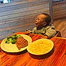 bowl, broccoli, child, clothing, dining, food, happy, hoodie, indoor, laughter, macaroni_and_cheese, meal, person, plate, restaurant, smile, steak, table, toddler, wooden_table