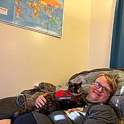 Gigi is registered to the contest to win money with this photo: blanket, blue_curtain, casual_clothing, cat, couch, glasses, hand, home, indoor, map, person, relaxing, remote_control, resting, service_dog_vest, short_hair, smiling, technology_device, tortoiseshell_cat, wall