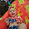Amara joined the competition — help win amazing prizes! baby, infant, unicorn_horn, headband, smile, happy, sitting, baby_chair, floral_pattern, colorful, clothing, red, white, blue, stars, stripes, indoor, furniture, living_room, person