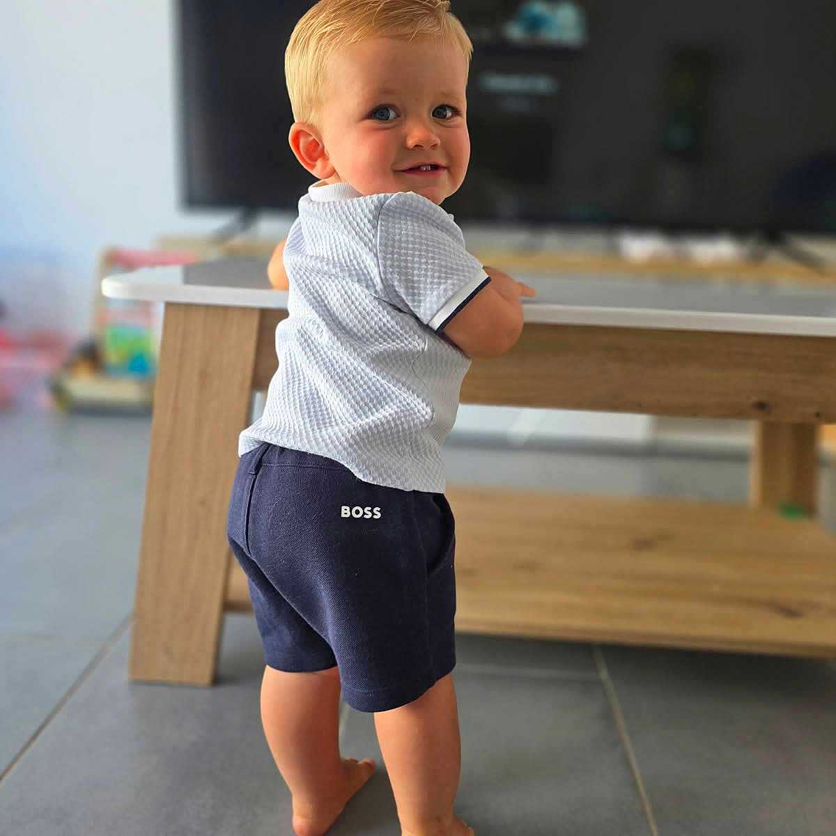 Tao participe au concours pour gagner de l'argent avec cette photo : baby, barefoot, blond_hair, casual_clothing, child, cute, floor, home, indoor, looking_back, person, portrait, shorts, smiling, standing, table, tile_floor, toddler, white_shirt, wooden_furniture