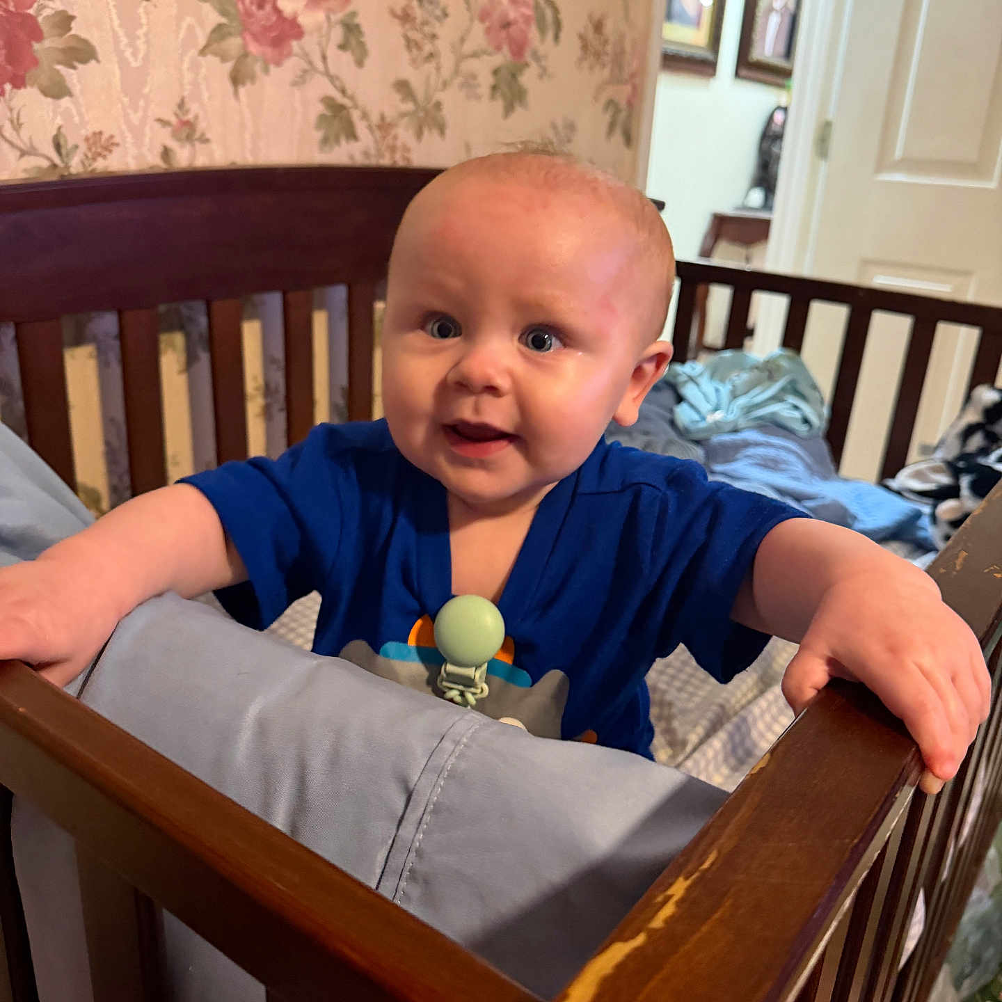 Liam is registered to the contest to win money with this photo: baby, crib, wooden_crib, blue_clothing, pacifier, infant, child, indoor, wallpaper, floral_pattern, furniture, portrait, smiling, grip, home, room, decor, person, young_child, curious