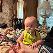 Liam is registered to the contest to win money with this photo: baby, infant, pacifier, yellow_clothing, tattoo, hand, bed, quilt, blanket, arm, furniture, wardrobe, lamp, vintage, curtains, room, indoor, person, holding, portrait