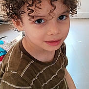 Nidal a rejoint le concours — aidez-le/la à gagner de superbes lots ! child, curly_hair, brown_shirt, indoor, portrait, smiling, young, person, window, natural_light, face, eyes, hair, casual_clothing, childhood, cute, looking_up, seated, home, bright
