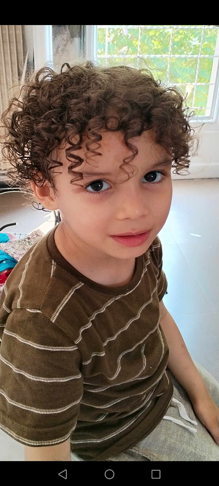 Nidal a rejoint le concours — aidez-le/la à gagner de superbes lots ! child, curly_hair, brown_shirt, indoor, portrait, smiling, young, person, window, natural_light, face, eyes, hair, casual_clothing, childhood, cute, looking_up, seated, home, bright