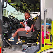 Sky Murdock joined the competition — help win amazing prizes! black_hair, child, clothing, dashboard, daylight, fan, fire_truck, hand, metal_floor, outdoor, person, pink_overall, reflection, seat, seatbelt, shoe, smile, steering_wheel, vehicle_interior, window