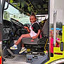 black_hair, child, clothing, dashboard, daylight, fan, fire_truck, hand, metal_floor, outdoor, person, pink_overall, reflection, seat, seatbelt, shoe, smile, steering_wheel, vehicle_interior, window