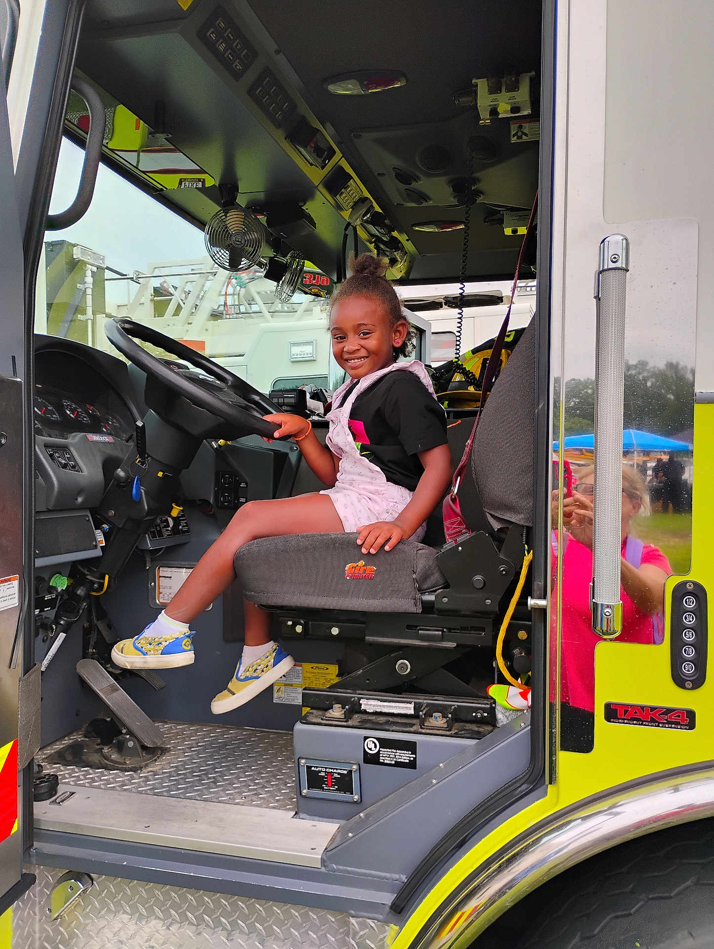 Sky Murdock joined the competition — help win amazing prizes! black_hair, child, clothing, dashboard, daylight, fan, fire_truck, hand, metal_floor, outdoor, person, pink_overall, reflection, seat, seatbelt, shoe, smile, steering_wheel, vehicle_interior, window