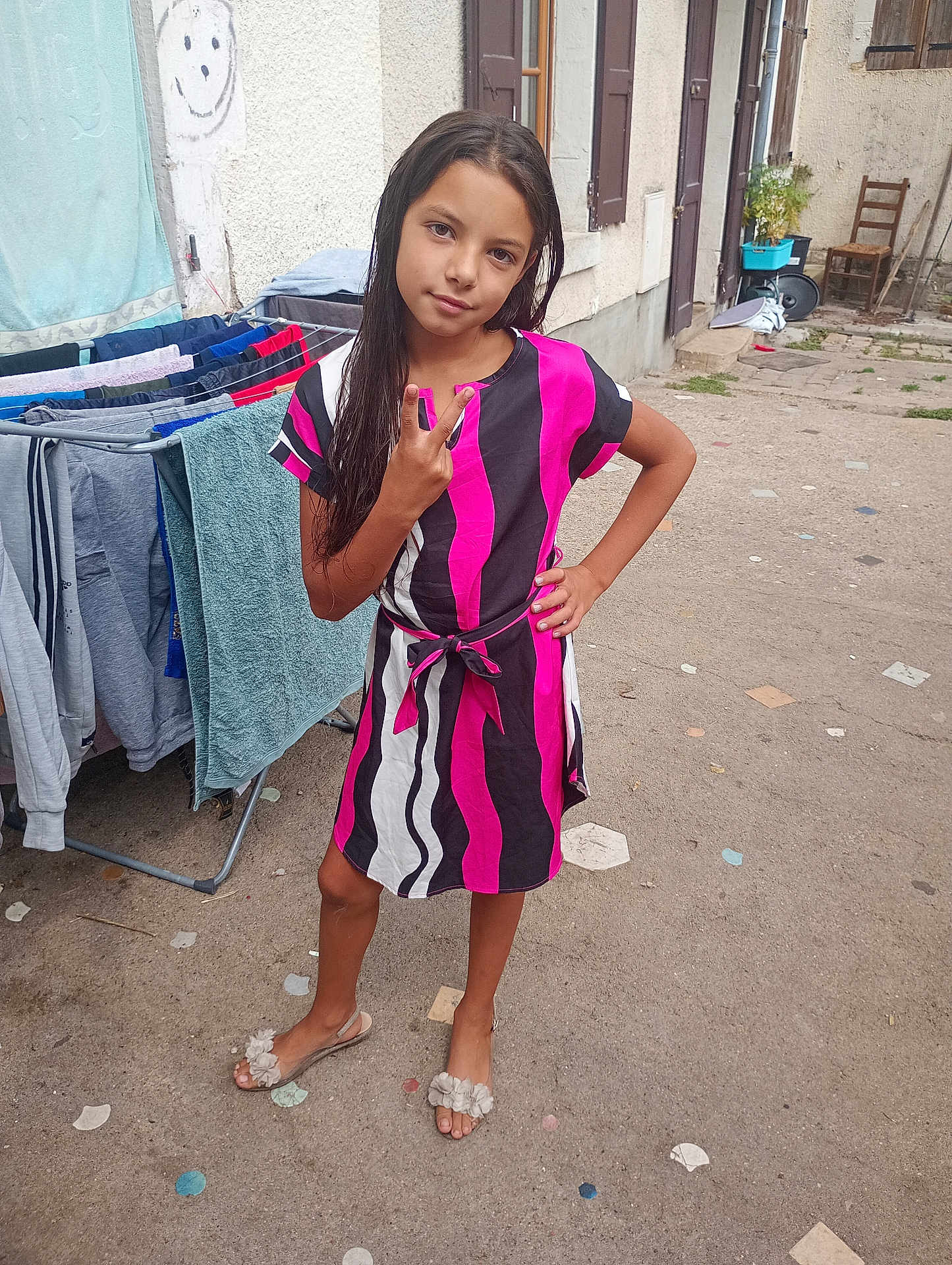 Naissy participe au concours pour gagner de l'argent avec cette photo : child, girl, dress, peace_sign, outdoor, laundry, drying_rack, smiley_face, concrete, footwear, hand_on_hip, long_hair, patterned_clothing, wall, window, door, chair, plant, casual, portrait