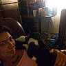 Diamond is registered to the contest to win money with this photo: person, cat, indoor, dim_lighting, reclining, chair, table, clutter, refrigerator, black_and_white_cat, hand, casual, relaxed, domestic, living_room, furniture, bottle, cup, background, light