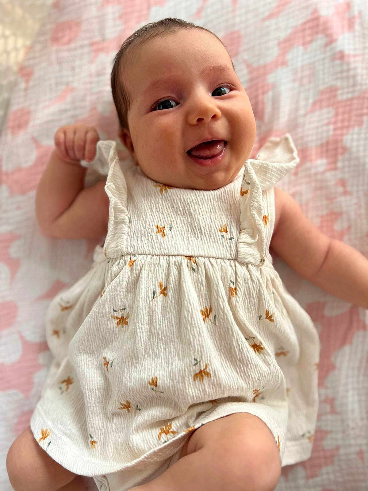 Alice participe au concours pour gagner de l'argent avec cette photo : baby, smiling, infant, dress, white_clothing, floral_pattern, blanket, pink, lying_down, tongue_out, cute, happy, child, skin, face, arm, leg, person, indoors, closeup