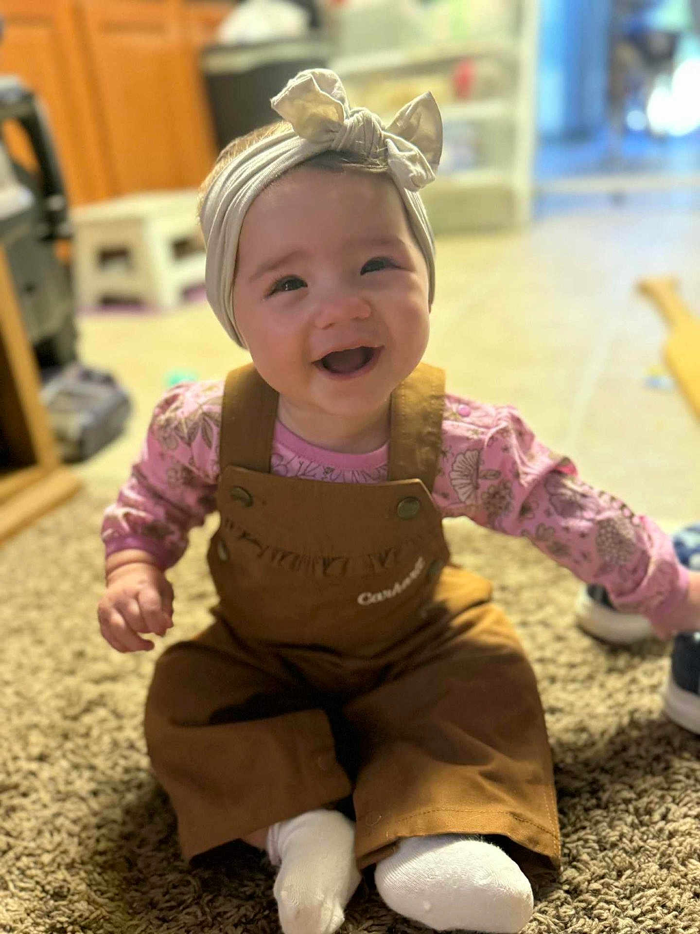 Amaya is registered to the contest to win money with this photo: baby, child, smiling, headband, overalls, sitting, carpet, indoor, happy, cute, clothing, footwear, infant, portrait, person, toddler, pink, brown, white_socks, home