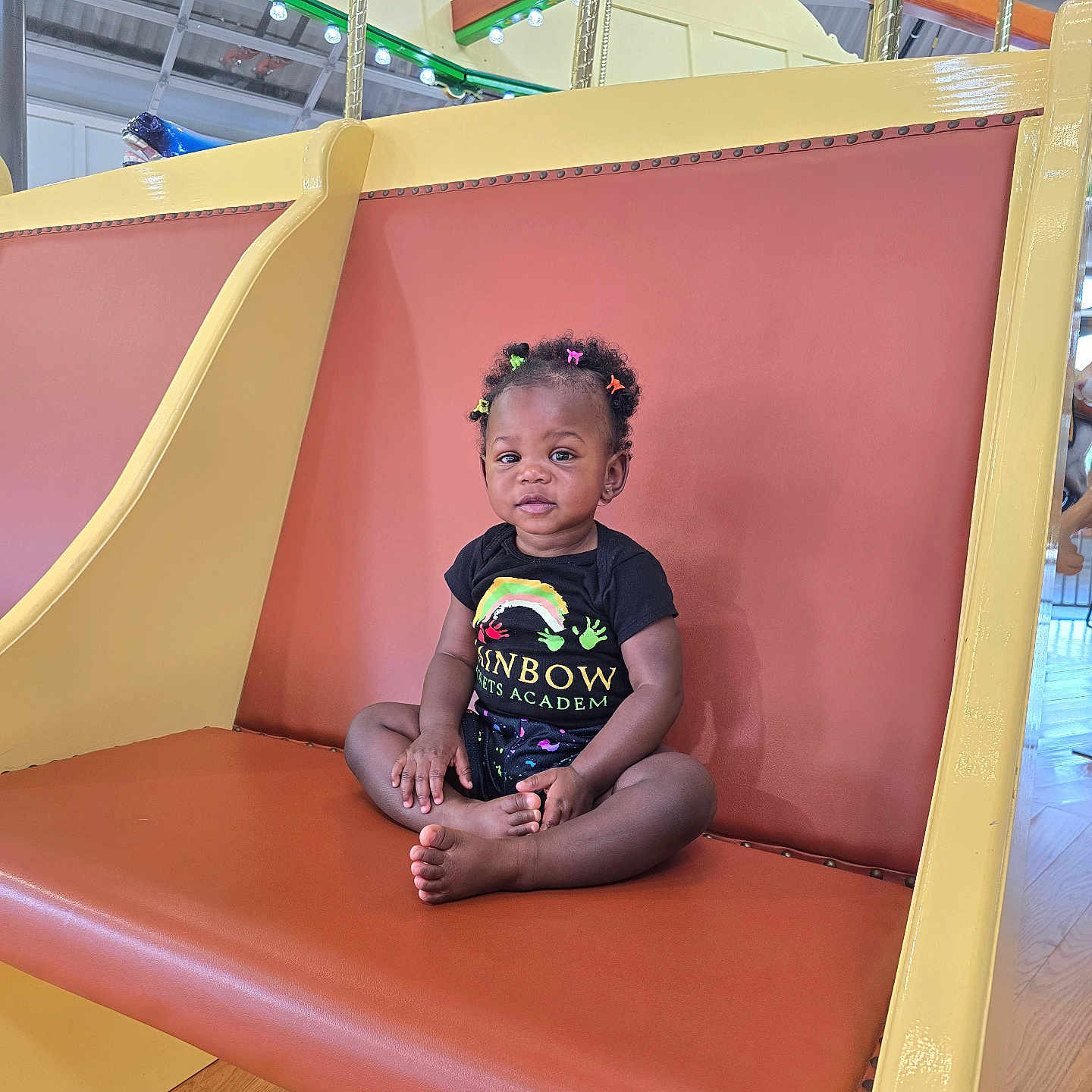 Naomi is registered to the contest to win money with this photo: black_shirt, bright, casual_clothing, child, colorful, cute, furniture, hair_clips, indoor, person, play_area, portrait, rainbow, seat, sitting, smiling, swing, toddler, wood_floor, young_child