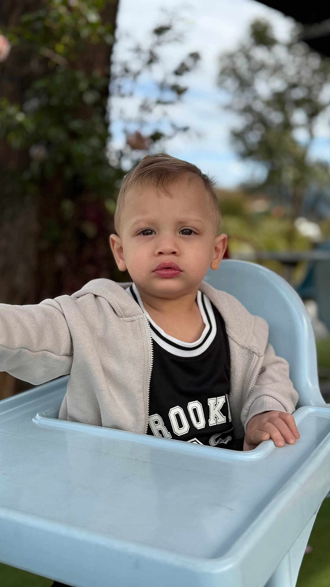 Kairo joined the competition — help win amazing prizes! toddler, child, high_chair, outdoor, hoodie, black_shirt, face, expression, hand, tree, sky, nature, seat, person, young_child, casual_clothing, daylight, portrait, serious, baby