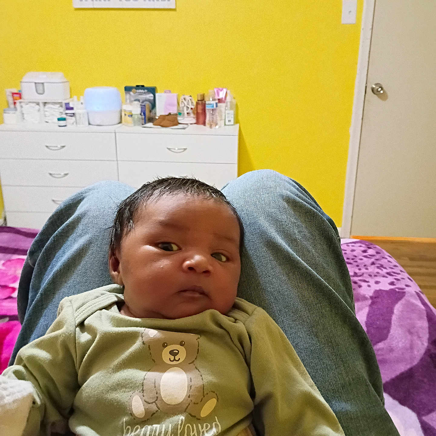 Oliver Smith is registered to the contest to win money with this photo: baby, infant, person, clothing, teddy_bear, green_shirt, jeans, lap, bedroom, yellow_wall, drawer, door, purple_blanket, wooden_floor, baby_essentials, face, hair, indoor, sitting, relaxed